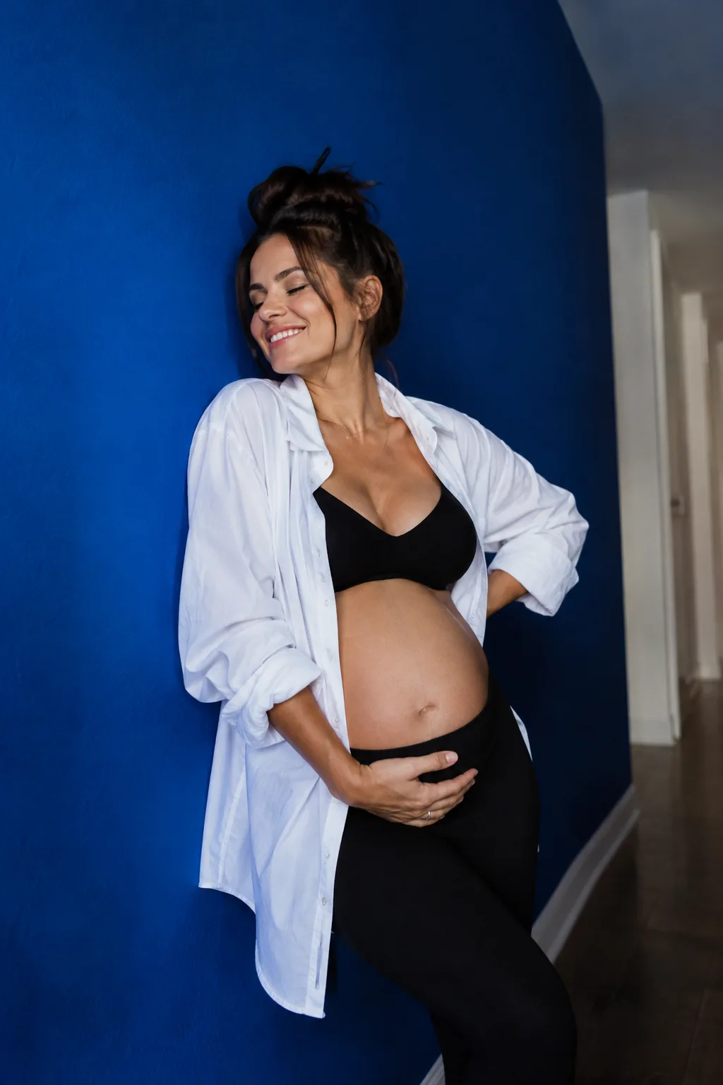 Pregnant woman in open button-down shirt leaning on hallway wall